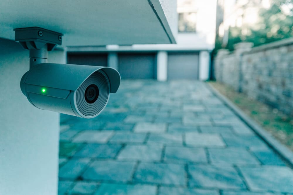 An exterior security camera mounted under a soffit, aimed down a home's driveway approach.