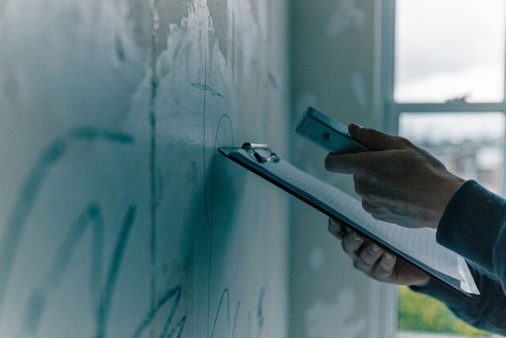 A hand holding a clipboard and laser measure against a bare interior wall during a site walkthrough.