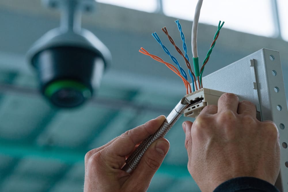A hand terminating a network cable into a keystone jack, with a ceiling-mounted camera behind.